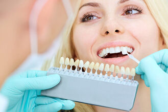 A dental professional in blue gloves holds a shade guide beside the patient's teeth, comparing color samples for veneers or crowns.
