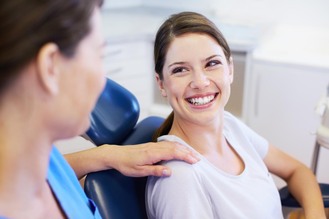 Patient seated in dental chair wearing white shirt while professional in blue uniform rests hand on shoulder in clinic setting.