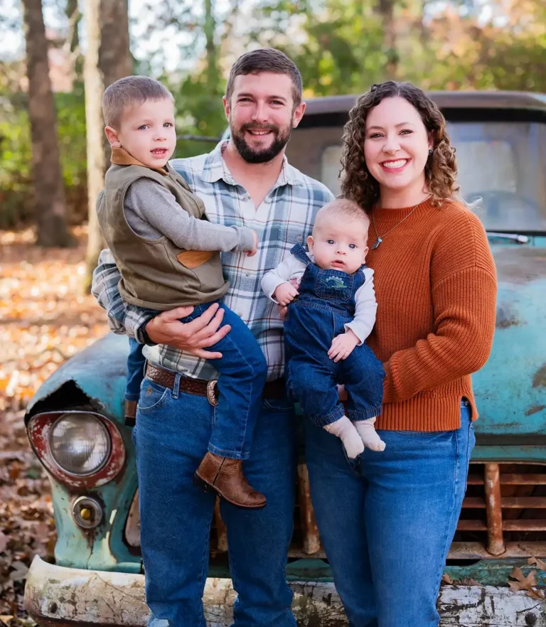 Family portrait outdoors in autumn with two adults standing in front of weathered turquoise car, each holding a child, surrounded by trees and fallen leaves.